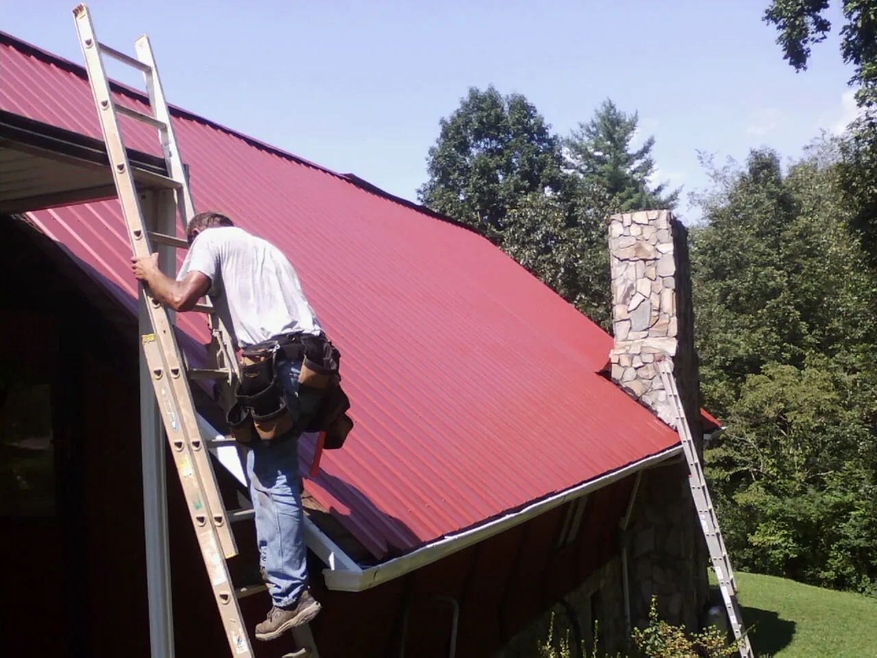 Roofer on a ladder beside a red metal roof and stone chimney — Soffit & Fascia Repair by Roofing Brothers in Ruckersville